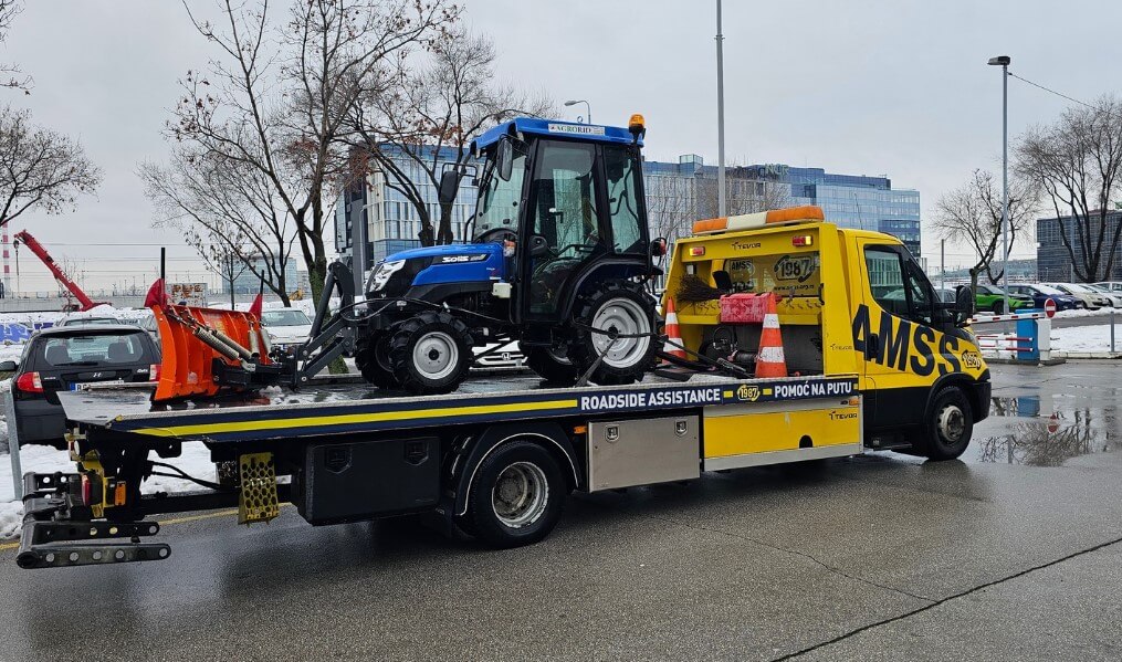 Šlep vozilo prevozi plavi traktor sa snežnim plugom